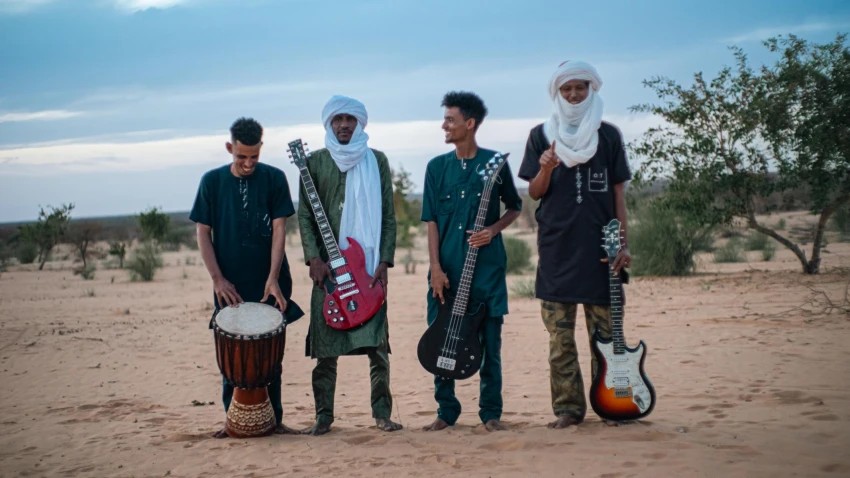 Four musicians with guitars and a djembe stand side by side in the desert at dusk