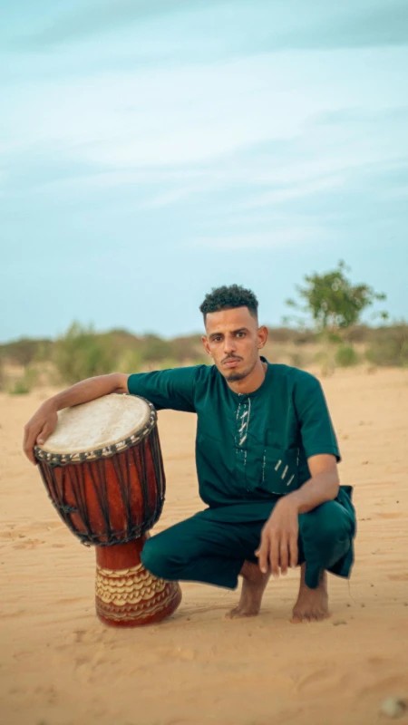 A musician in a green tunic crouches in the sand next to a djembe