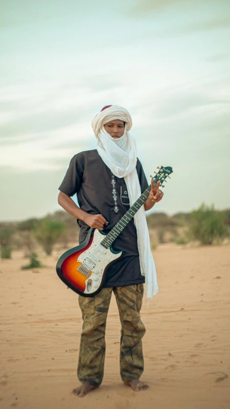 A young musician in a white turban stands barefoot in the desert with an electric guitar