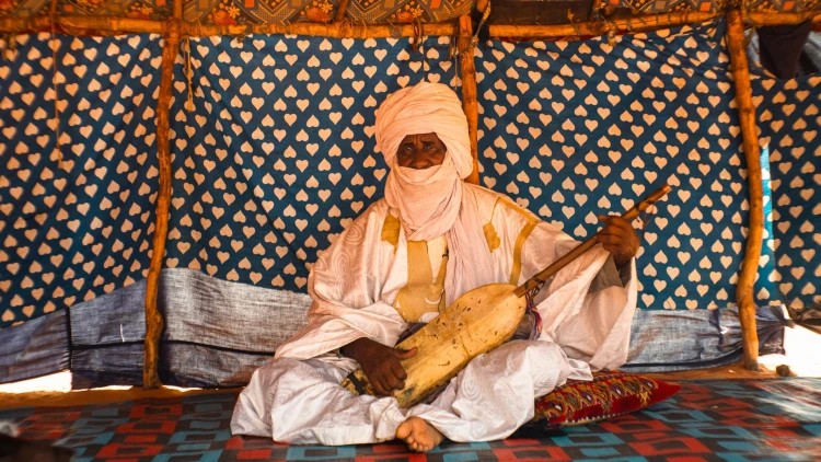 An older musician in white robes sits in a patterned tent holding a traditional lute
