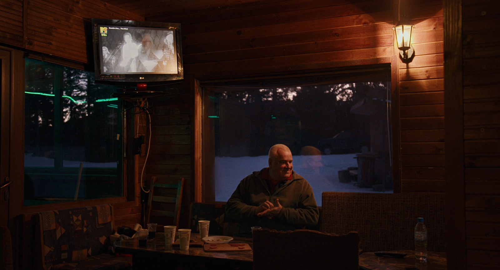 A man sits at a table in a wood-panelled room, a television on the wall behind him