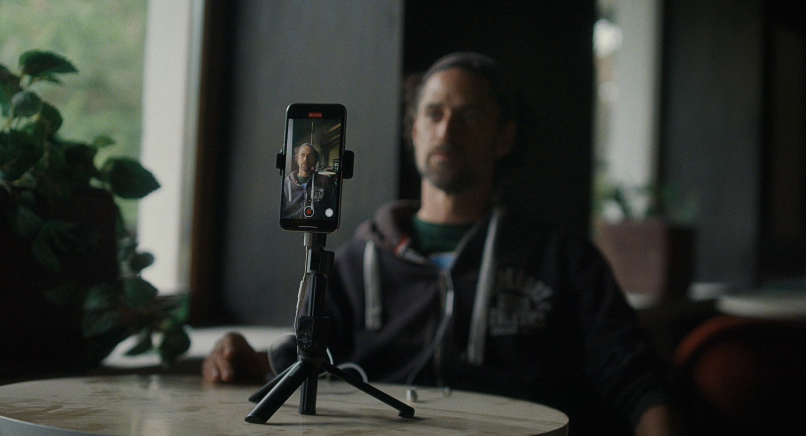 A man sits behind a table while a smartphone on a tripod films him
