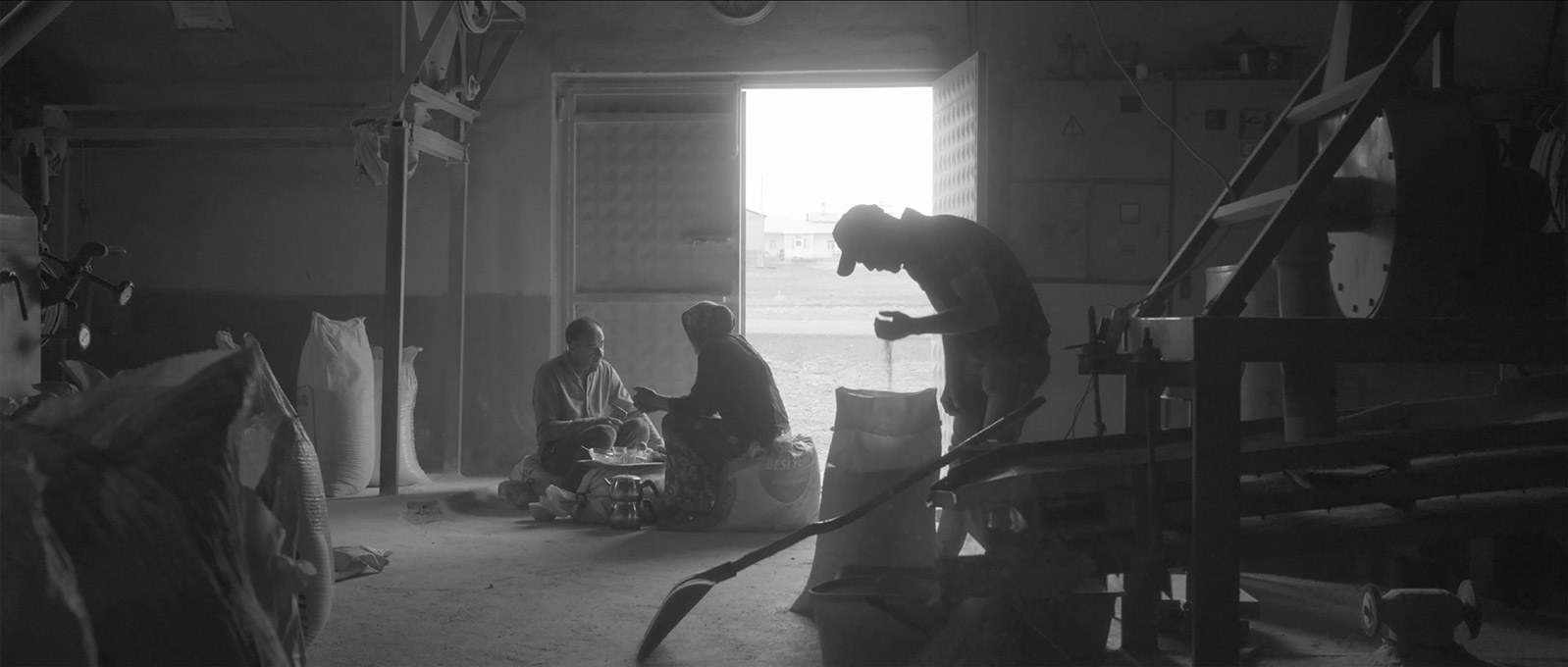 Two people share tea on grain sacks in a storehouse while a man lets seeds run through his hand