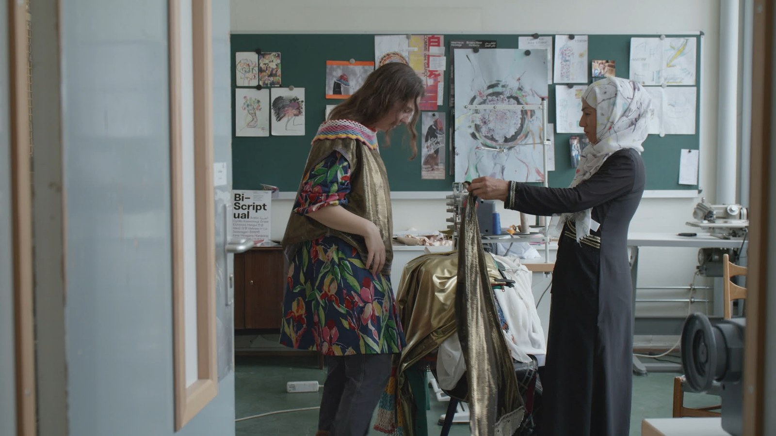 Two women fit a garment in a sewing workshop with drawings pinned to a board