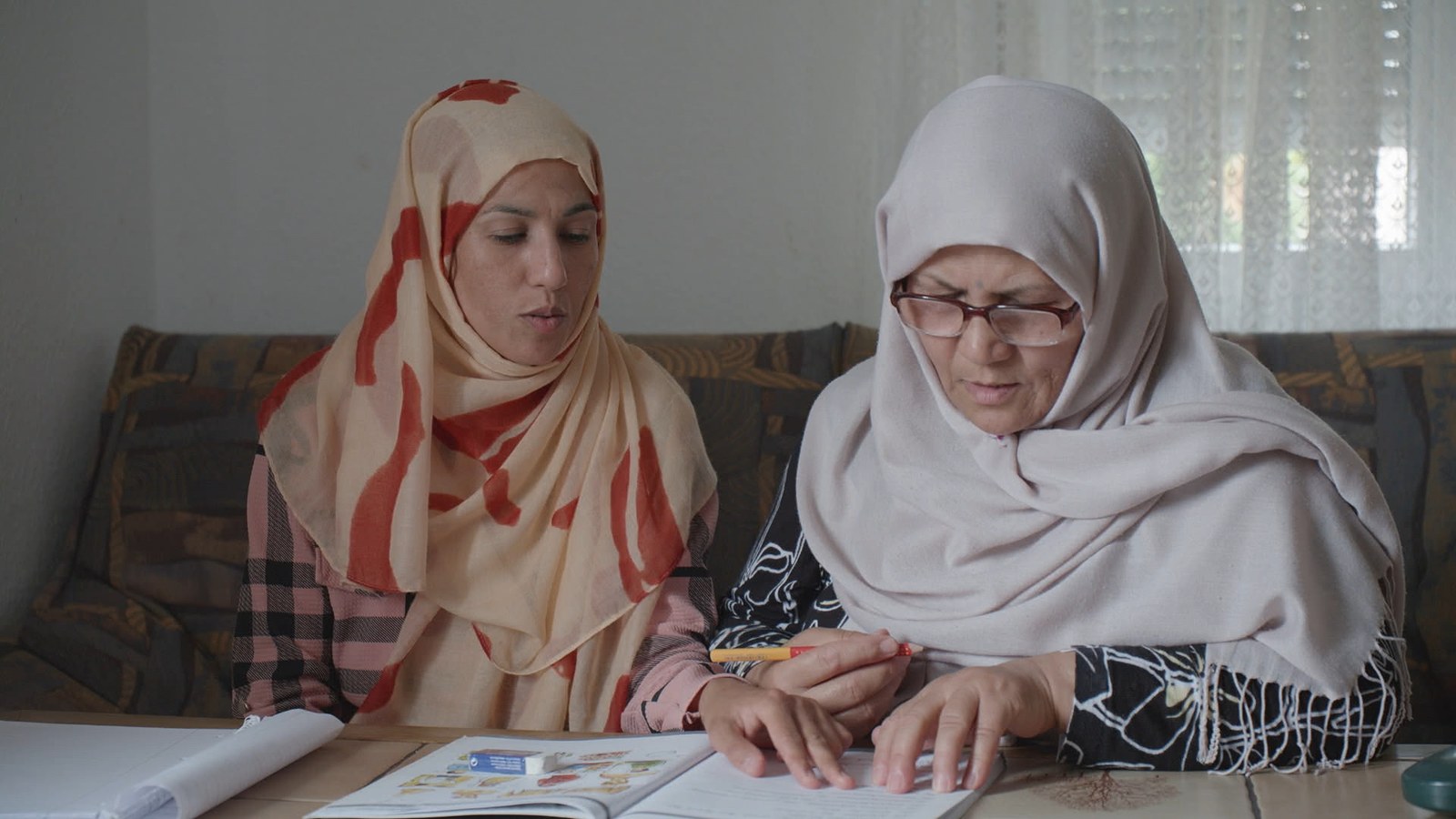 Two women in headscarves sit on a sofa and work through a language exercise book