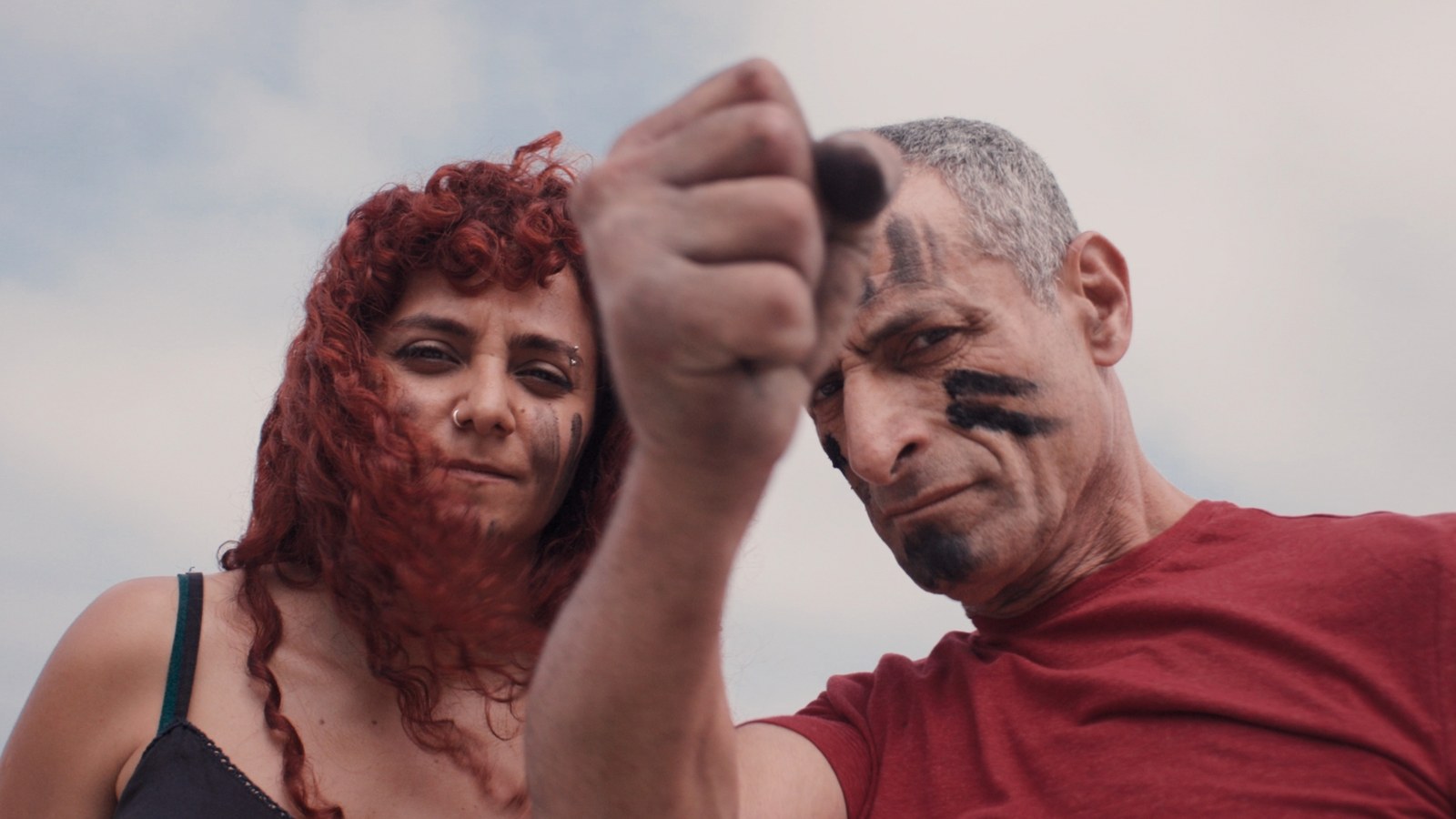 A woman and a man with black paint on their faces look into the camera, his fist in front of the lens