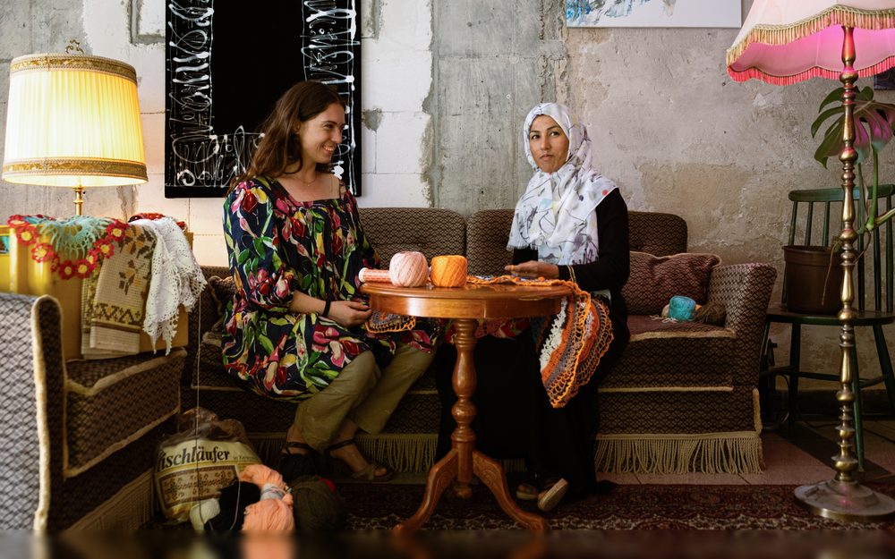 Two women sit on an old sofa at a small round table with balls of wool